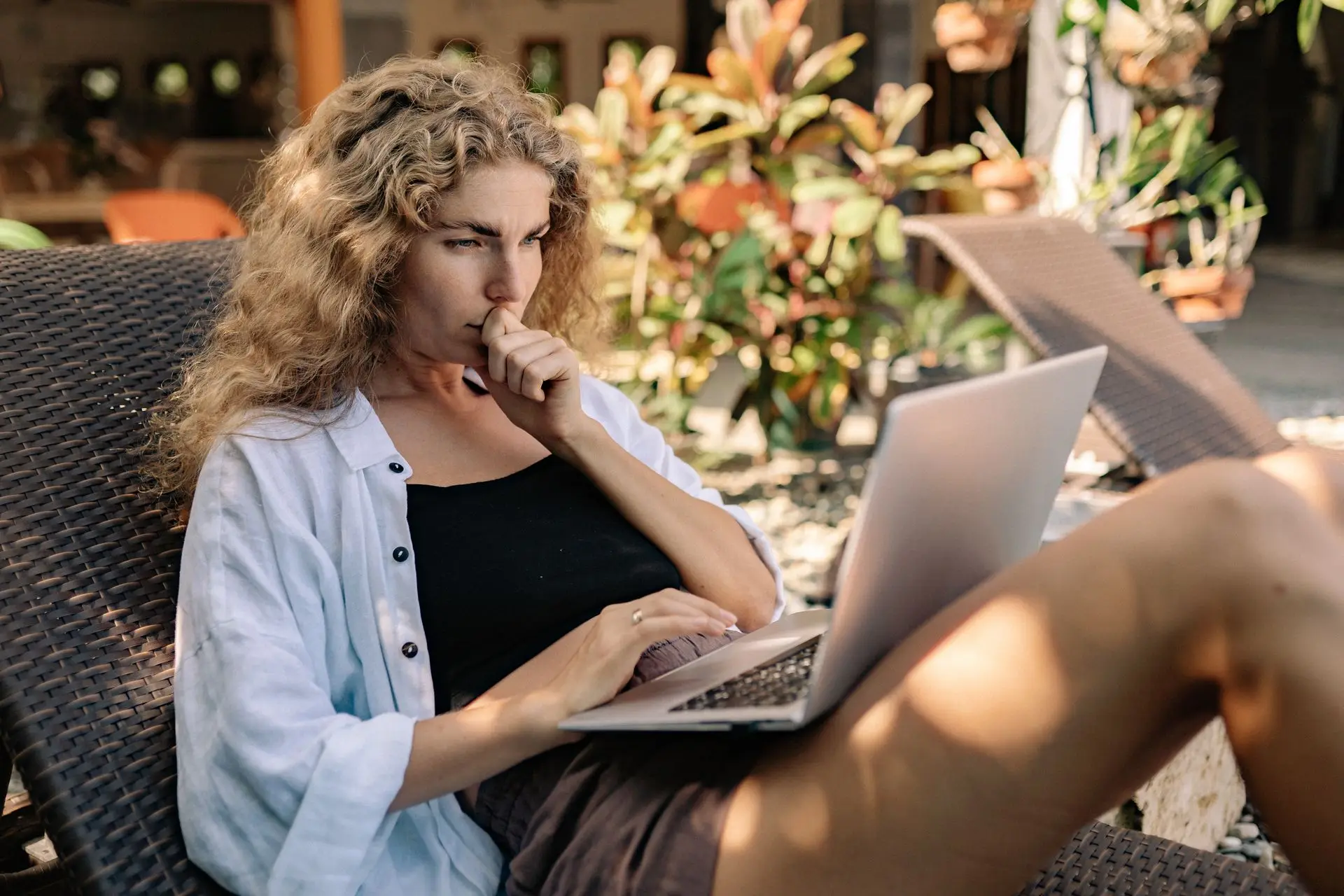 Woman working on a laptop from a sunlit patio lounger surrounded by tropical plants