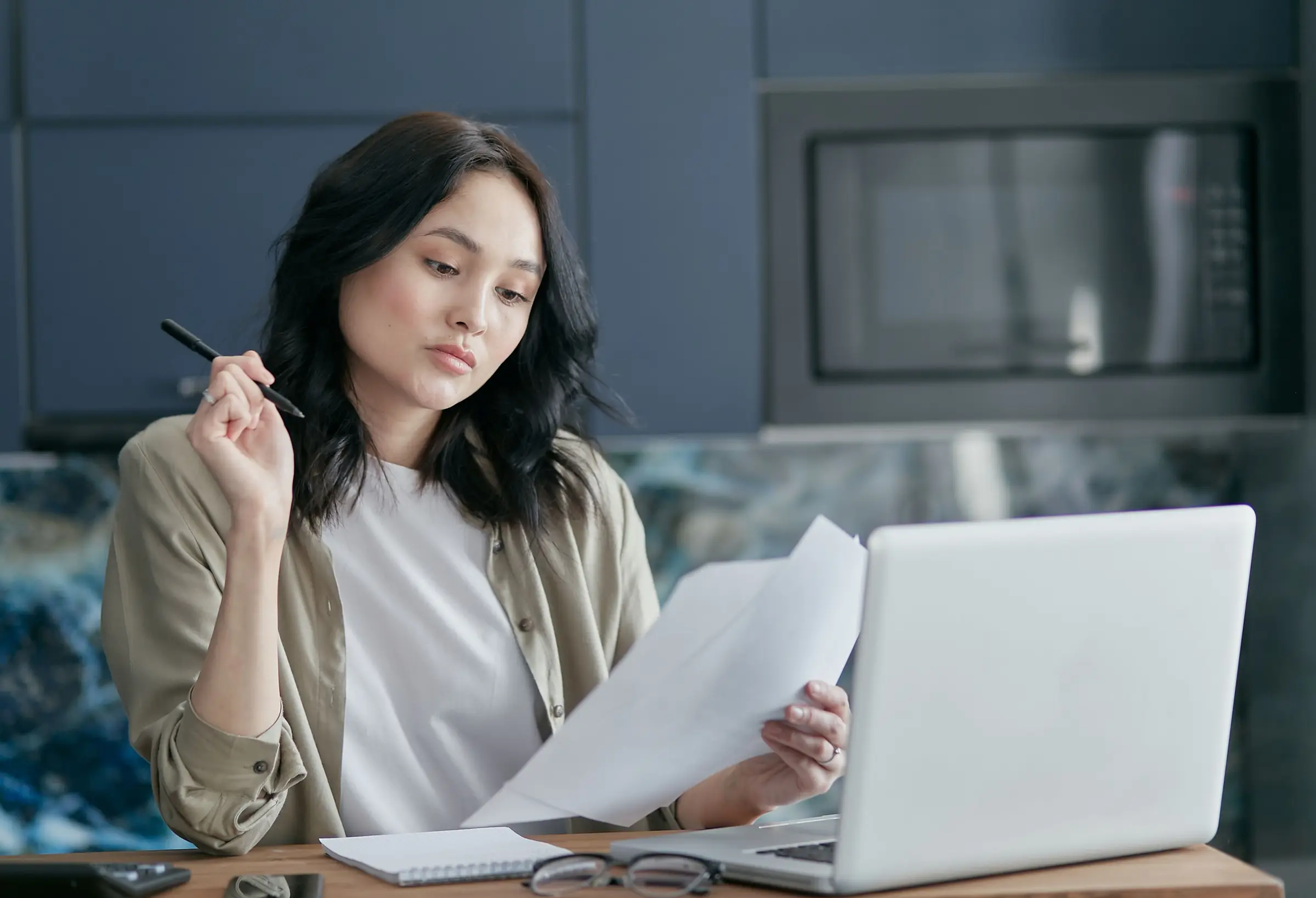 Woman at a kitchen table holding a pen, reading a printed policy page beside an open laptop