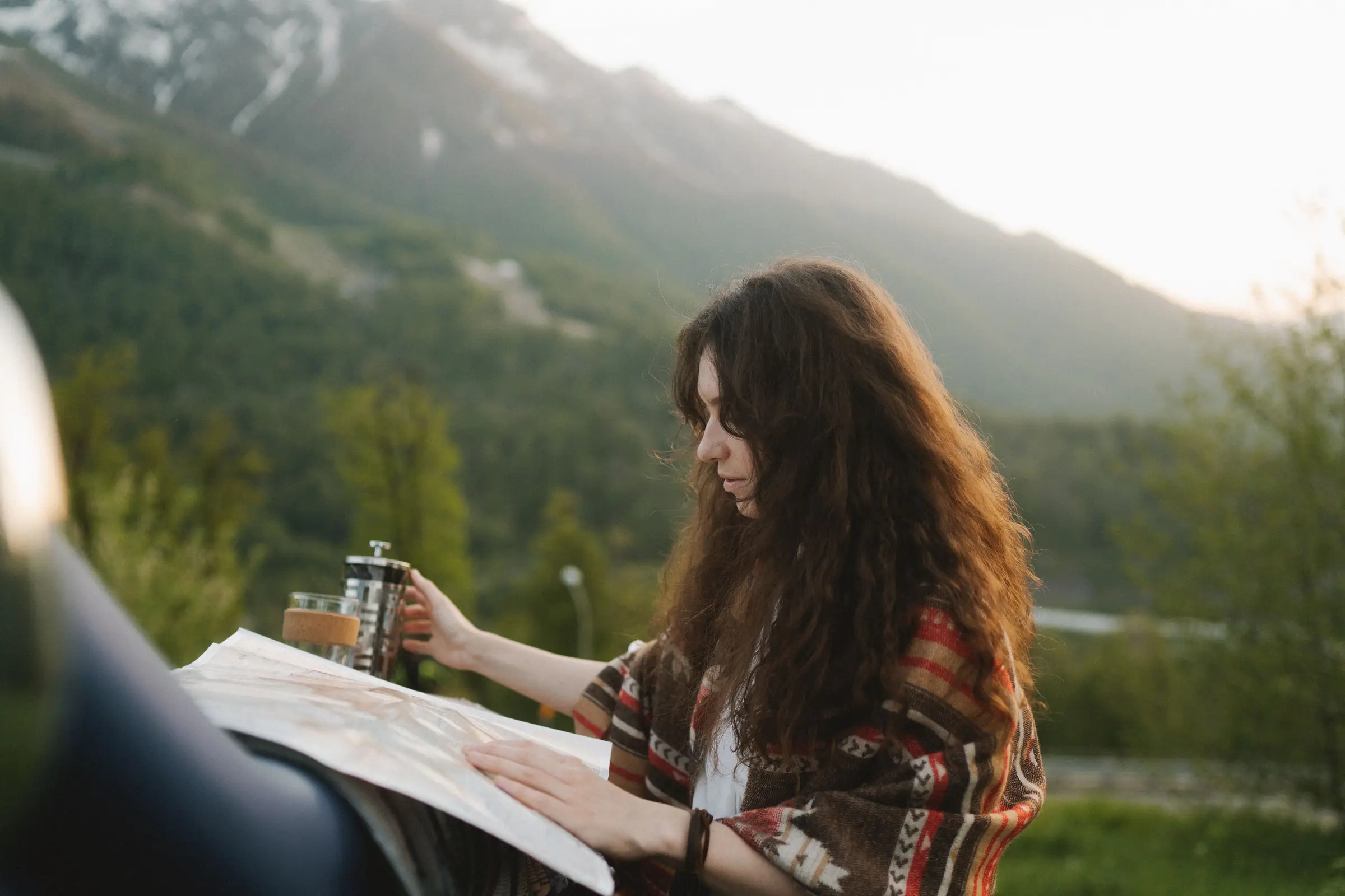 Woman reading a paper map spread on the hood of a car, with mountains behind her